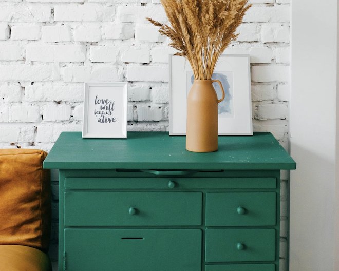 The green dresser near the breaks decorated wall. On the comode there are small and big white decorative white frames and wheat color vase with bouqet of spikes of the same color in it. On the left of the dresser there is a part of the pillow also of the wheat color. And the pillow in the wheat color on left of the dresser