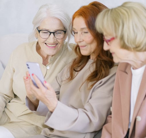 three white elder women are looking into a phone that the woman in the middle is holding